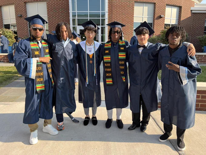 Six graduates wearing gowns and caps pose for a picture before graduation.