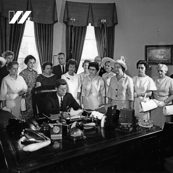 A square black and white image of President John F. Kennedy signing the Equal Pay Act of 1963 at his desk with a group of women and then-Vice President Lyndon B. Johnson standing behind him. There is a white “w” U S O W logo in the top left corner.
