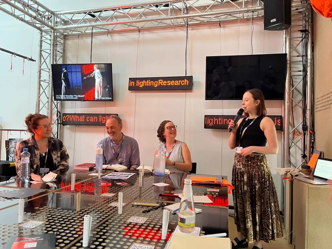 Four figures talking around a table with a map of UK . Screens on wall show examples of theatrical work and scrolling text provocations: What can light do? What is a Lighting Q? How does light affect space, time, action?