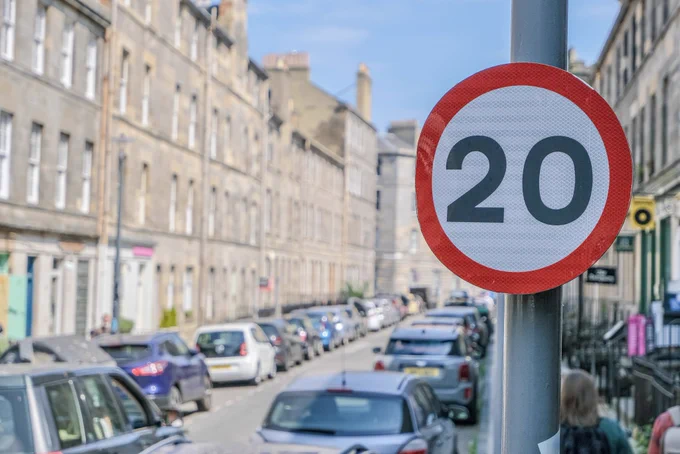 A close-up of a red and white 20mph speed limit sign in a residential neighborhood of Edinburgh lined with limestone buildings.