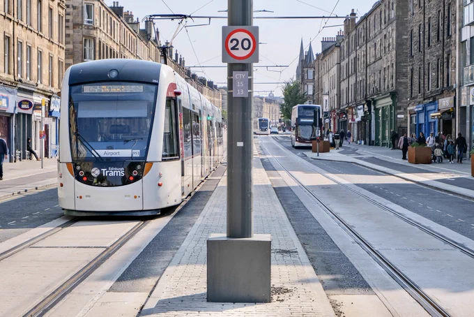 A modern white tram moves on a commercial shopping street in Leith, Scotland, a small 20 mph speed limit sign mounted to a post.