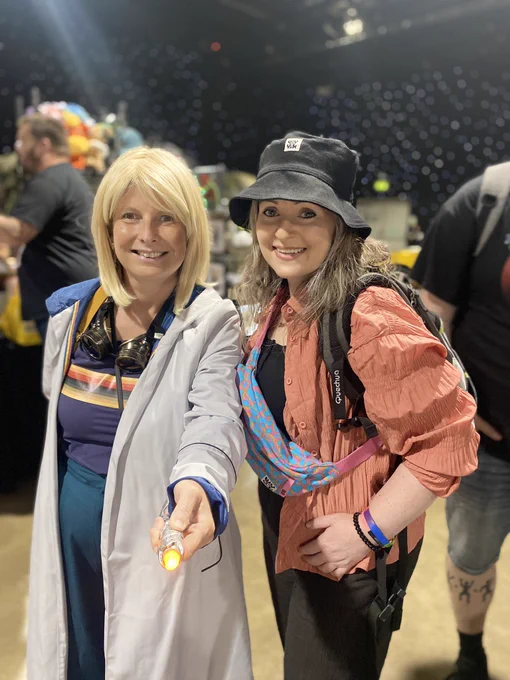 This is a photo of two women on the left is a woman dressed in a cosplay costume as Jodie Whittaker’s version of Doctor Who, so she has a blonde wig, hungry, anorak NAV T-shirt, with rainbow stripes horizontal across the chest and blue calotte trousers. She is holding a stainless steel version of the sonic screwdriver on the right is a white woman, wearing a black bucket hat, a coral pink over shirt and black top and bottoms. They are both smiling.
