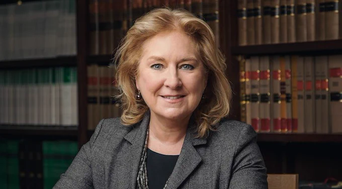 Photo of Dame Sue Carr, a smiling white woman with shoulder-length blonde hair, grey blazer an black top underneath. She is sitting in front of a bookshelf of legal texts.