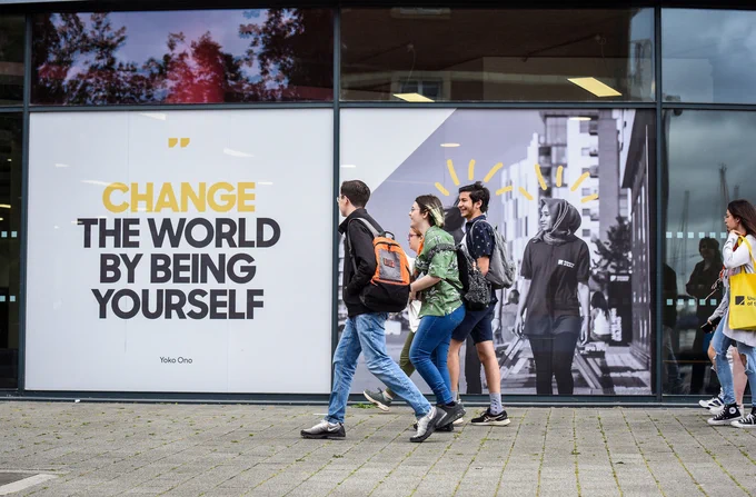 A group of students walk outside the front of the University of Suffolk, past a sign reading "Change the world by being yourself"