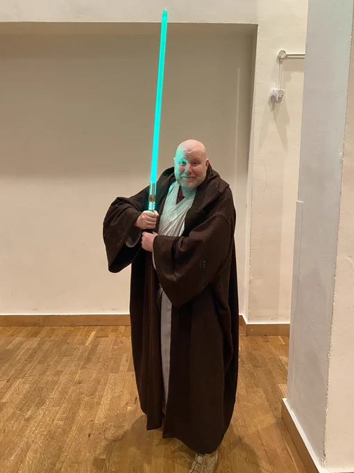 A tall white man who looks about 50 years old with a bald head, and a very short white beard and moustache he has a long brown Jedi cloak on with long beige undershirt, and he’s holding up a huge green light sabre