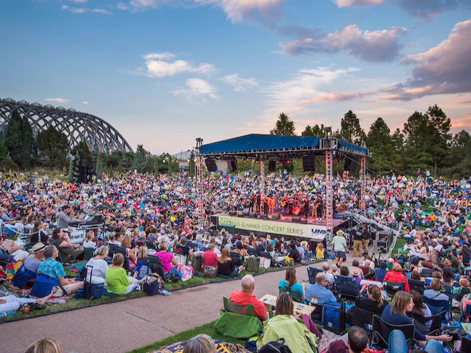 A lot of people sit on the amphitheater grass surrounding a tall square stage. A row of trees lines the horizon with large clouds in a clear sky. The conservatory sits in the background.
