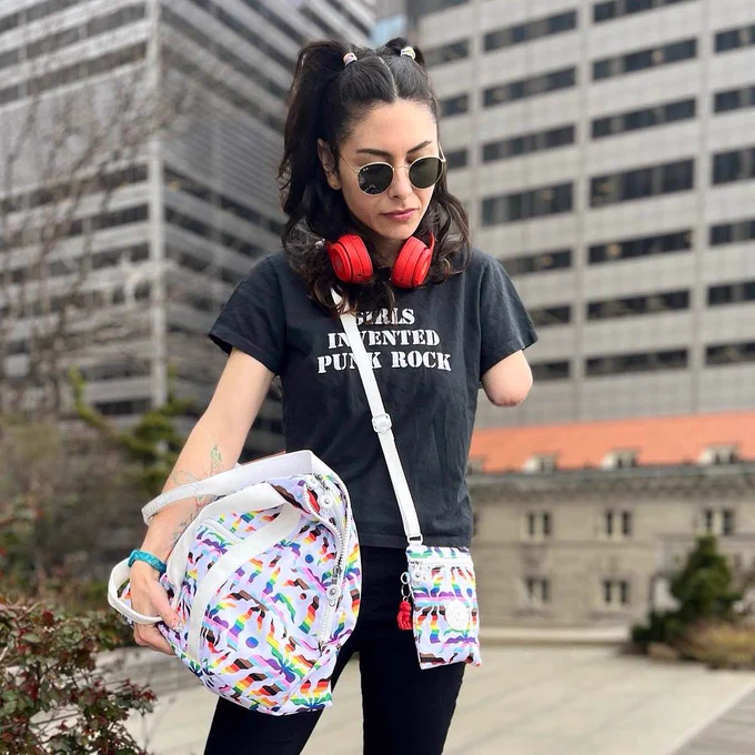 Photo Description: Chloé is wearing black skinny jeans and a black tee with a graphic that reads “girls invented punk rock.” She’s pictured from the knees up and glancing down. She’s carrying a rainbow Kipling duffel in her right hand and has a rainbow Kipling crossbody pouch on her left side. She also has on cherry red headphones around her neck and round Ray Bans.