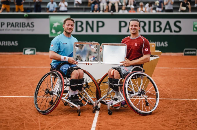 Image shows Alfie Hewett (left) - Alfie is a white male with short brown hair. He is in his sports wheelchair wearing a pale blue t-shirt, holding a silver plaque and smiling. Gordon Reid is on the right. Gordon is a white male with long brown hair pulled back in a bun, he is wearing a red t-shirt, holding a silver plaque and smiling. They are situated on the tennis court which is dark orange with white lines. 