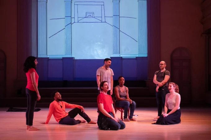 A group of people singing together wearing red, white, and grey clothing. Some sit casually or posed, others stand. They all face different directions. A minimal black and white drawing of a receding road is projected behind them.