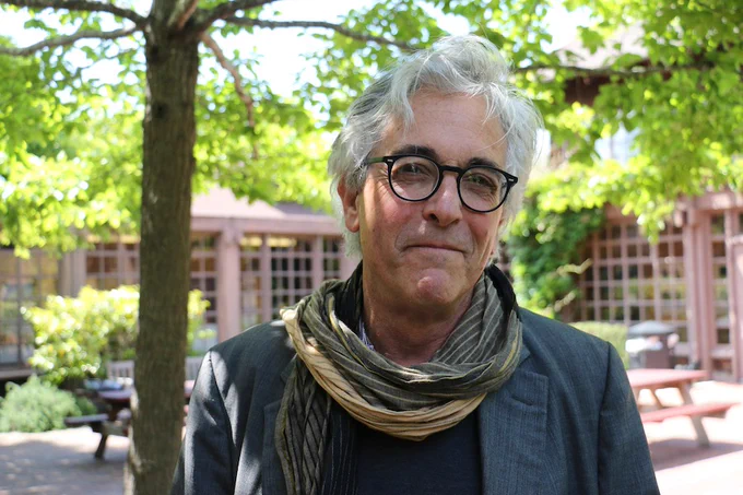 Photo of award-winning investigative reporter Mark Schapiro wearing black glasses with a striped scarf over a gray suit jacket in the North Gate Hall courtyard with trees in the background.
