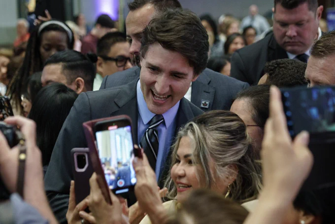 Prime Minister Justin Trudeau is standing in a crowd of people and posing for a photo with a woman. Both of them are smiling. The woman is holding her cellphone up to take the photo, and other cellphones are visible.