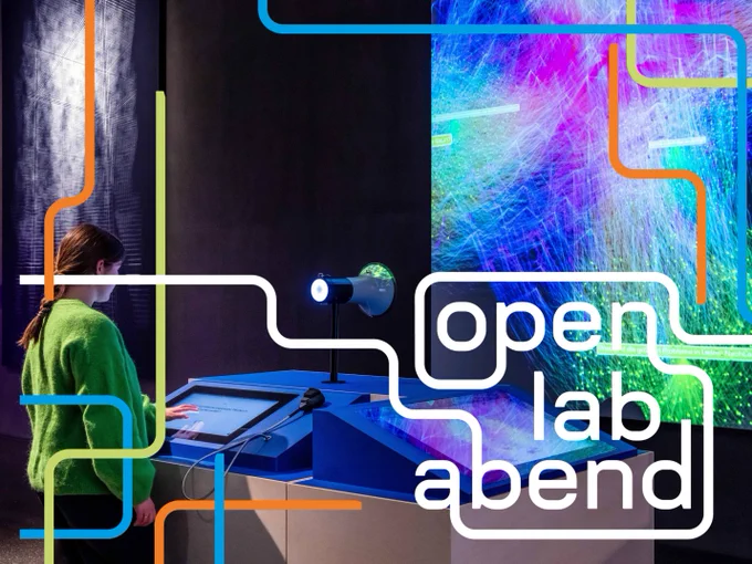 Lab im Futurium. Hinten rechts ein großer Bildschirm, auf dem viele bunte Vernetzungen zu sehen sind. Davor mittig das Exponat "Data Correlations" mit zwei Bildschirmen sowie einem Megafon und Mikrofon. Links daneben eine Person, die den Bildschirm berührt. Darüber das OLA-Keyvisual: Orange, grüne und blaue Rohre zieren das Bild an den Seiten. Aus weißen Rohren entsteht die Schrift „open lab abend“.