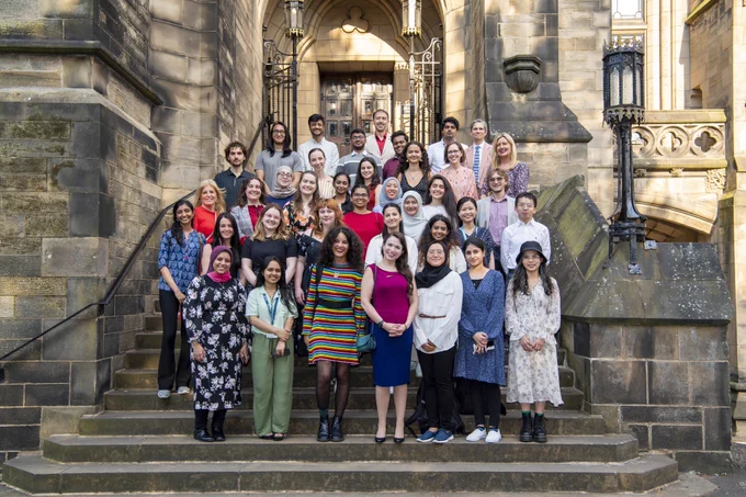 A group of people (men and women) standing on the steps at the University of glasgow chapel with iron gates in the background and light catching the right hand side of the building. The group are all smiling at the camera.