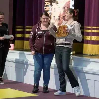 three women present their idea on stage. One is holding a wooden object and pointing to it
