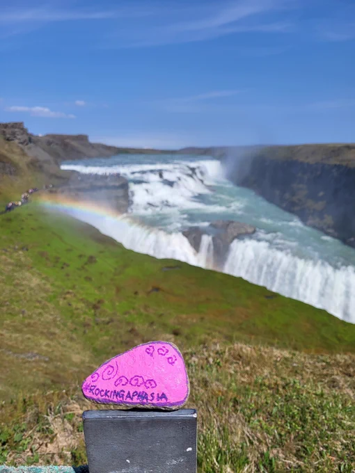 Photographe with a pink and purple painted #RockingAphasia stone in  and an icelandic waterfall with rainbow spray in the background. The Sky is clear and blue