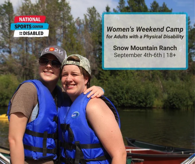Two women are in life jackets on the river. One woman has her arm around the other and they are both smiling. 