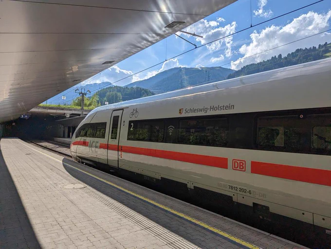 Ein ICE4 steht am Bahnsteig. Über dem Zug sieht man grüne Berge und blauen Himmel