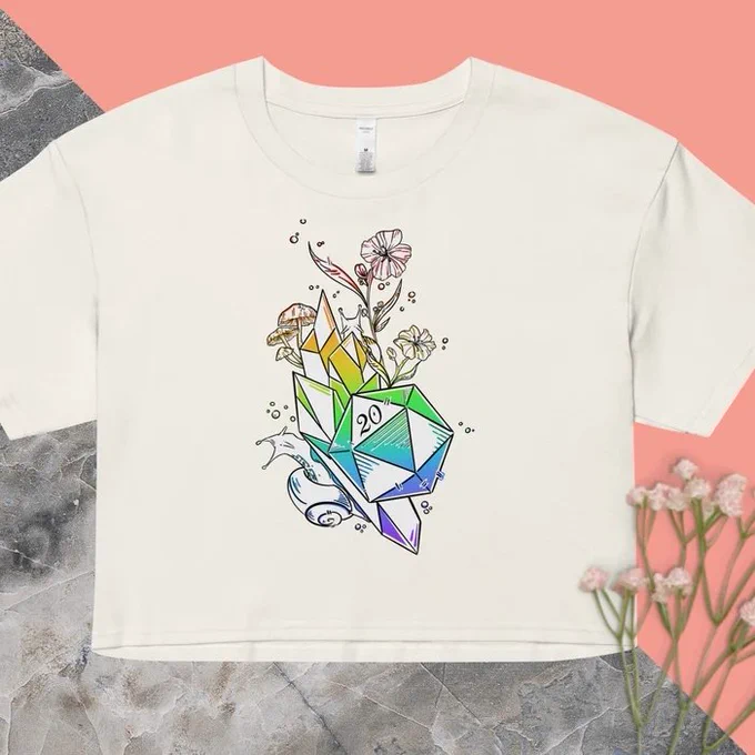 A photo of a cream colored t shirt on a grey marble and salmon pink background. The shirt had a rainbow colored graphic featuring a d20, crystals, mushrooms, flowers, and super cute snails!