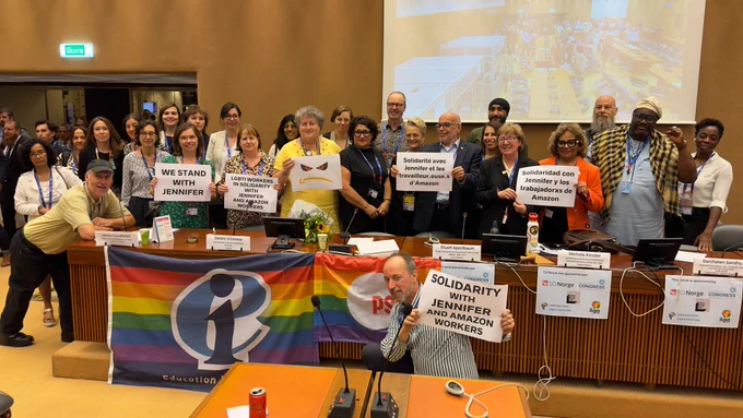 A group of people stand behind a podium holding "We Stand with Jennifer" and "Solidarity with Jennifer and Amazon Workers" signs in multiple languages.
