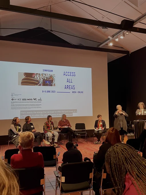 A seated audience watches five seated presenters, one standing presenter and one interpreter in front of a large projected screen. The screen shows an image that is mostly white that includes small black text and large blue text that reads “Access all areas"