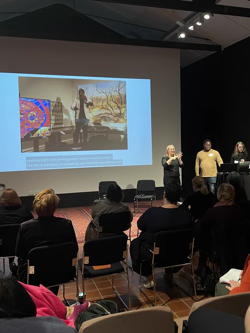A seated audience watches two presenters and an interpreter stand in front of a large projected screen. The screen shows an image of one of the presenters standing in an art gallery. Below the image are captions of the presenter's speech.