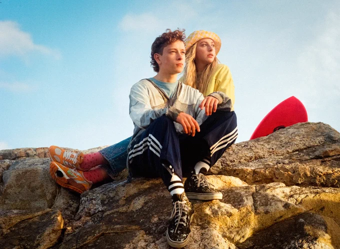 A young man and woman sit on a rock bathed in sunlight, both looking towards something out of frame. Part of a surfboard can be seen behind them.