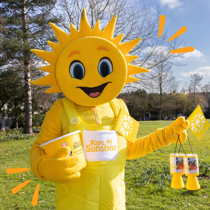 A mascot with a sun head holding Ray For A Day flags and fundraising buckets.
