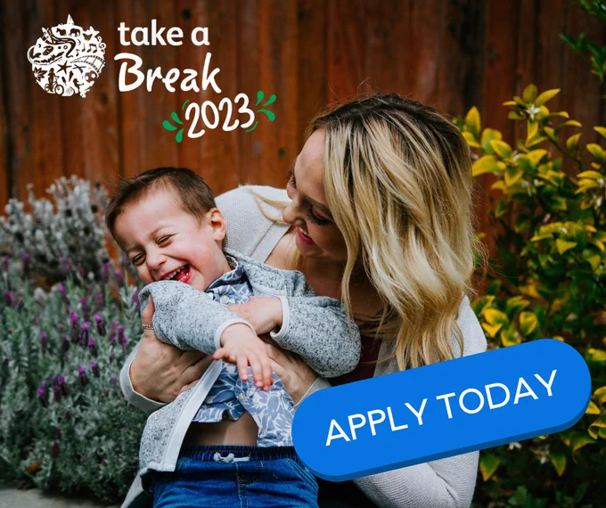 A blonde haired lady cuddles a young child she has in her arms. They are both smiling. They're in front of a fence with flowers and plants. The Take a Break 2023 logo sits in the top left. In the bottom right is a button sitting at an angle that says Apply Today.