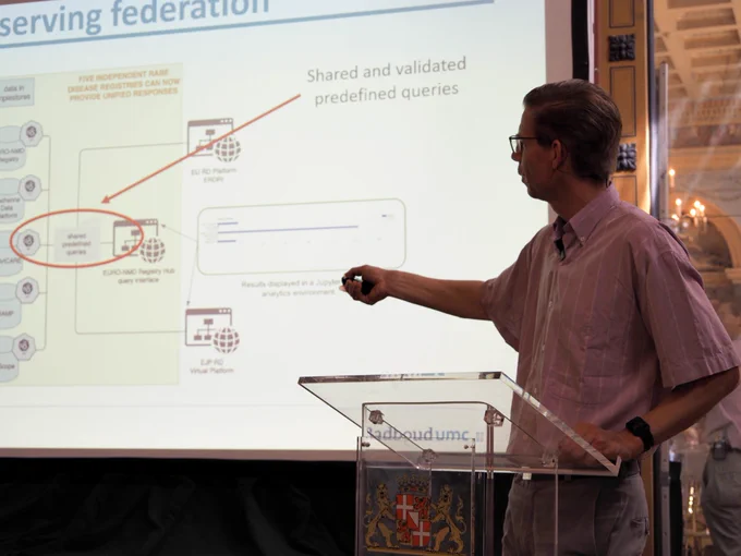 A white man wearing glasses and a summer shirt points a clicker at a presentation