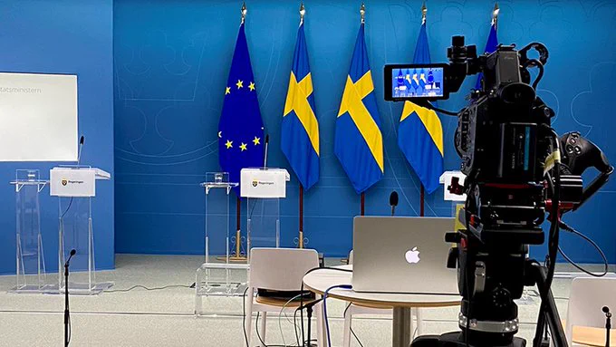 Bild på pressträfflokal. talarstolar står uppradade framför flaggor, kamerautrustning i förgrunden. 