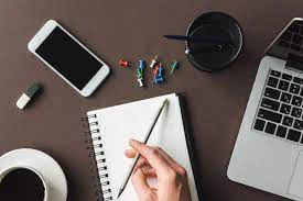 A stock image of a desk. The desk has a cup of coffee, some thumbtacks, a mobile phone, and a laptop. Someone is holding a pen above a notebook.