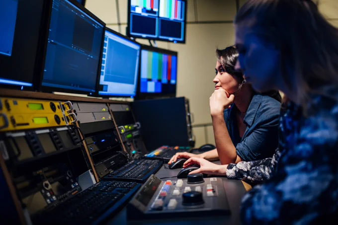Two people sit in front of monitor screens and editing equipment  
