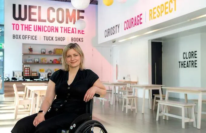 Rachel Bagshaw smiling at the camera in the Unicorn Theatre foyer. She has short blonde hair and is wearing all black. Large text on the wall behind her reads 'Welcome to the Unicorn Theatre, Box Office, Tuck Shop, Books'
