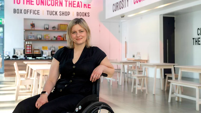 A white woman dressed in black clothes sat on a wheelchair, smiling. There are chairs and tables behind her. At the back there is a display of colourful items and colourful writing on the wall.