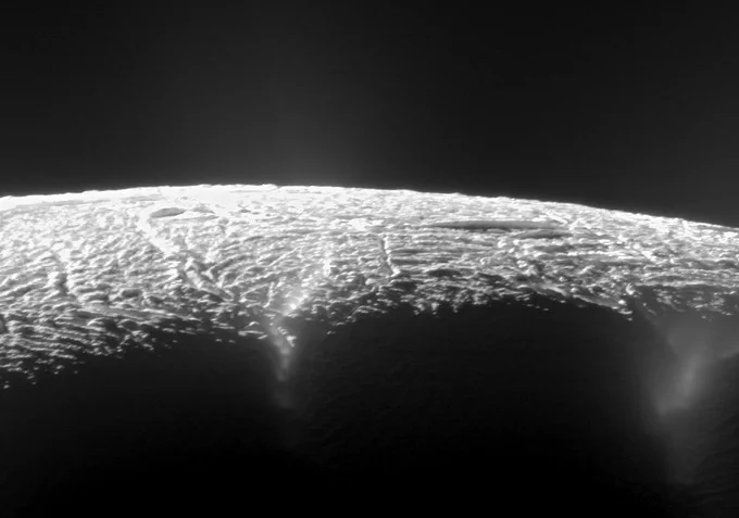 The curving horizon of Saturn's moon Enceladus as seen from space by the Cassini spacecraft. The surface has a rough, wrinkled texture. From fissures in the surface, misty plumes are seen rising.