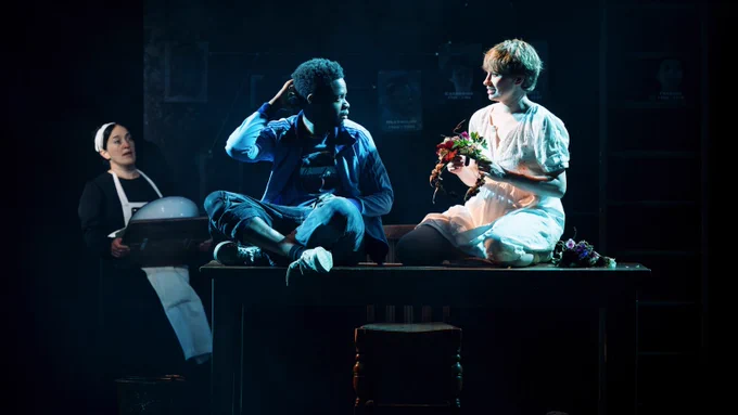 Two people sit on top of a table under a spotlight. One of them is holding a flowery head dress. Behind them is a woman dressed as a house keeper.