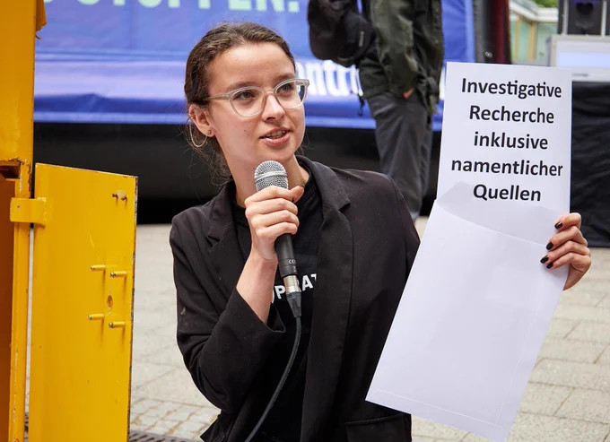 Anne hält neben dem offenen Briefkasten einen geöffneten Brief, auf dem "Investigative Recherche inklusive namentlicher Quellen" steht. 