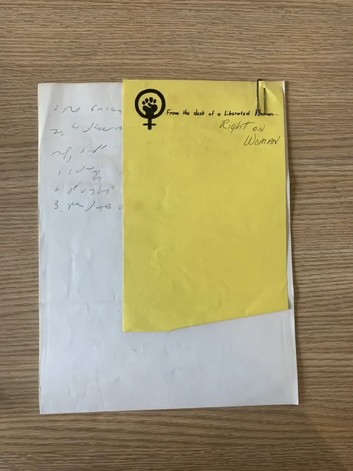 a white piece of paper with handwritten notes with a yellow paper paperclipped to it. The yellow paper has the women power fist icon and reads ‘from the desk of a Liberated Woman.’ Below that in handwriting reads, ‘Right on Woman.’ 