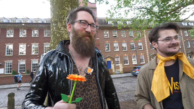 Tom Clark is stood outside the Yogscast Studios smiling in a leather coat holding a plastic flower. Tom is sporting a yellow ribbon around his neck smiling too.
