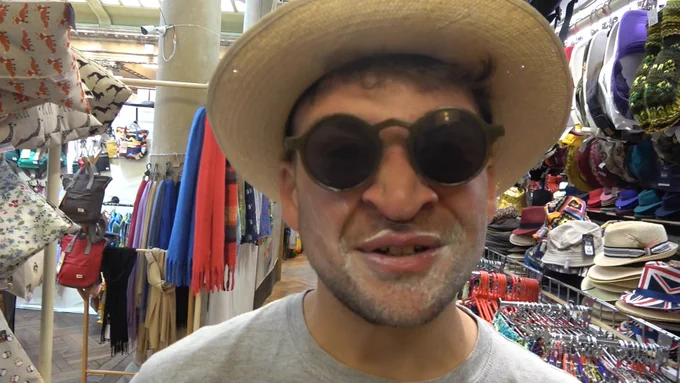 Daf is in Bristol Market surrounded by hats and gloves. He's wearing green round sunglasses, a straw hat and has sun cream smeared all over the bottom half of his face (he looks silly).