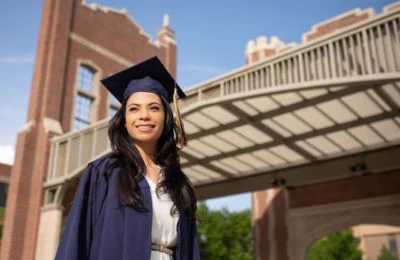 A young woman in a dress, graduation robe and graduation cap smiling. 