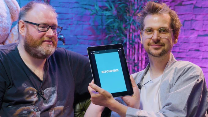 Simon & Lewis are sat on the peculiar portion set. Simon is looking displeased, Lewis is holding up an iPad with "Bitchfield" on the screen, he has a cheeky smirk on his face.