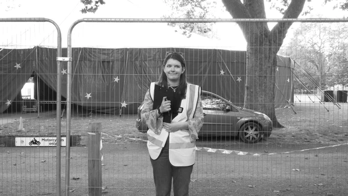 A black and white photo of Sarah in a high-vis jacket holding a clipboard. She's stood in front of a tall, metal fence that's blocking the entrance to a circus-like tent that's in the middle of Queen Square.