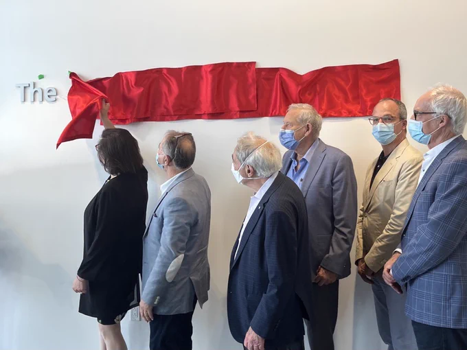 A group of people in business dress. One person is removing a red ribbon from the wall to reveal a sign.