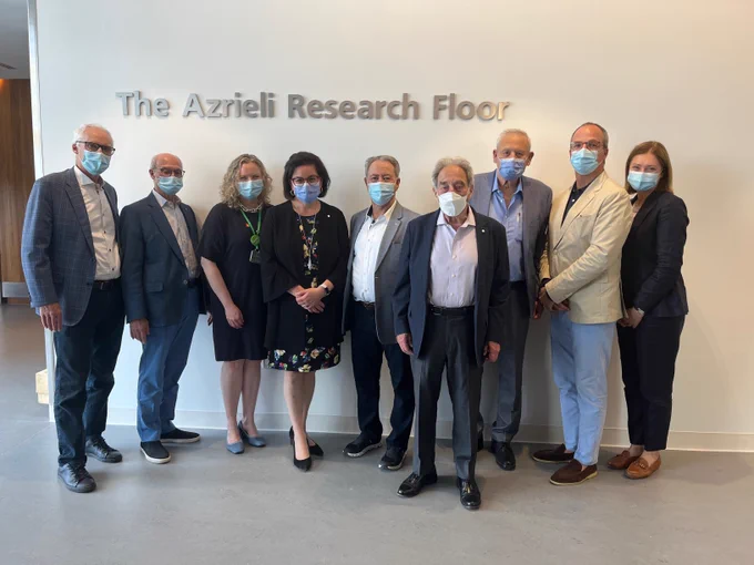 A group of people dressed in business-wear and masks standing in front of a sign that reads The Azrieli Research Floor.