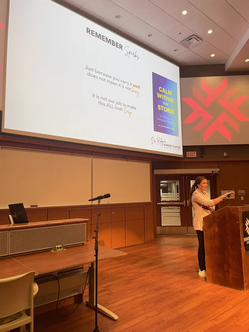 Dr. Robyne speaking at the podium at Fanshawe College in front of the slide: just because you carry it well does not mean it’s not heavy. It is not our job to make it all look easy.