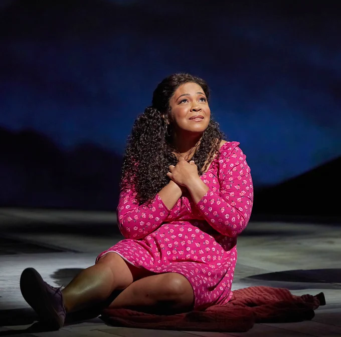 A woman in a pink dress sits with both her hands on her heart