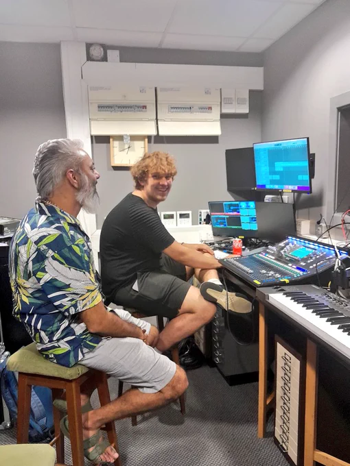 Loki and Oli are in a small room. They both sit at a table with screens, lighting and sound desk, and music keyboard. Loki looks at a screen. Oli turns to the camera and smiles.