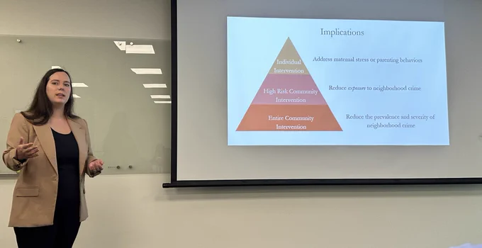 This photos is a candid photo of Rebecca Brady, a female PhD candidate with brown hair who is wearing beige blazer and black slacks, presenting her PhD dissertation. The slide shown depicts the implications of her study with a pyramid representing the levels of intervention. The bottom level reads “Entire community intervention - Reduce the prevalence and severity of neighborhood crime.” The next level of the pyramid reads: “High risk community intervention - Reduce exposure to neighborhood crime.” The top level of the pyramid reads: “Individual Intervention: Address maternal stress or parenting behaviors.”