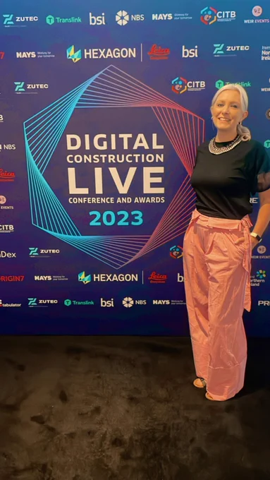 A blonde white woman with her hair in a ponytail. Wearing red baggy trousers and a black top. Her hands are in her pockets and she’s smiling. She is standing infront of a banner which is purple with various logic and in large white text Digital Construction Live conference and awards and 2023 in blue.